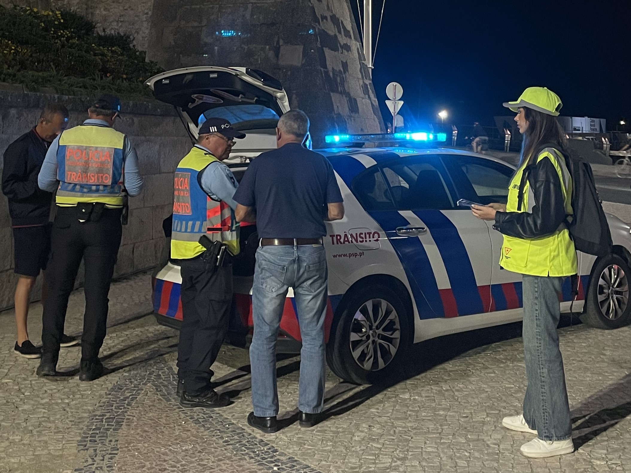 Agentes da pol&iacute;cia de tr&acirc;nsito abordam um condutor junto a uma viatura policial, durante uma opera&ccedil;&atilde;o de fiscaliza&ccedil;&atilde;o rodovi&aacute;ria, &agrave; noite. Um elemento da ANSR acompanha a opera&ccedil;&atilde;o.
